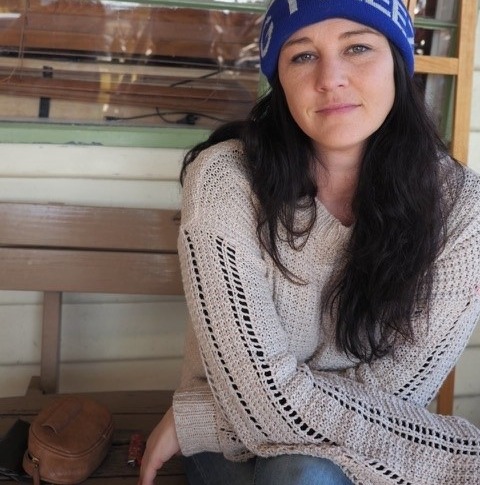 Image of a girl in her 20s or 30s sitting on a wooden bench. She's staring directly into the camera with a solemn expression and her arms crossed across her lap